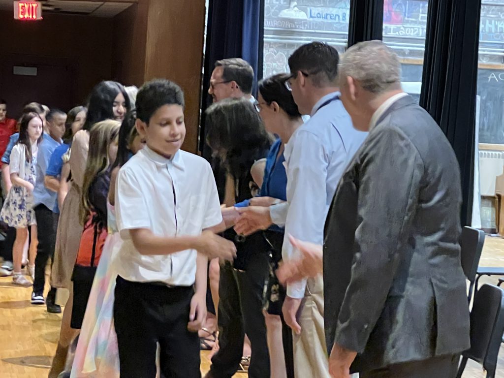 A young man with short dark hair, wearing a white shirt shakes hands with a man wearing a suit jacket.