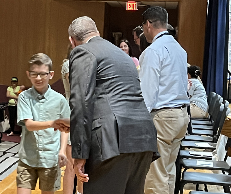 A fifth-grade boy with short dark hair, glasses and wearing a short-sleeve green shirt shakes hands with a man in a suit jacket.