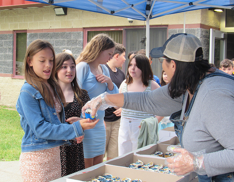 A group of fifth-grade girls stand at a table as a woman hands out cupcakes.