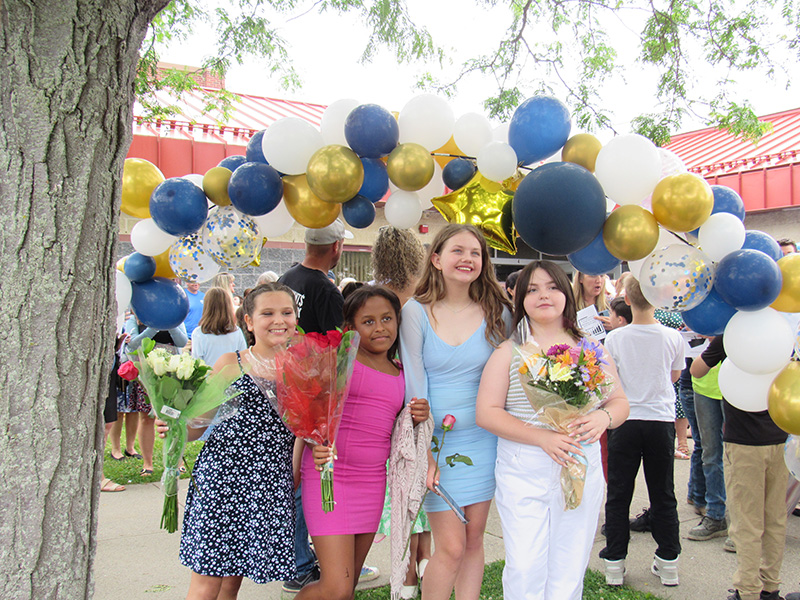 A group of four fifth-grade girls stand together, many holding flowers, under a balloon arch of blue and gold.