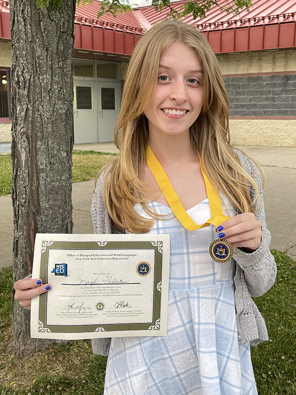 A young woman with long blonde hair holds her medallion in one hand and a certificate in another. She is smiling.