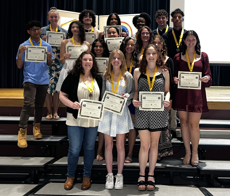 A group of 17 high school seniors on risers stand together holding certificates.