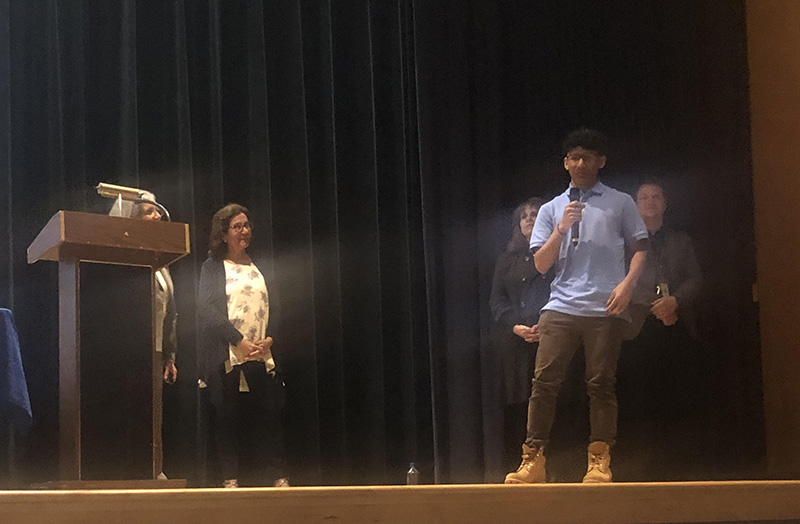 A young man, wearing a blue shirt and holding a microphone, talks while teachers around him are listening.