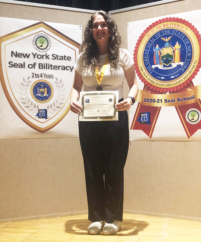 A young women with long dark hair holds a certificate. She is smiling and has a medallion around her neck.