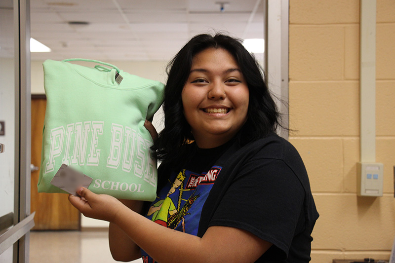 A woman with dark hair holds up a light green sweatshirt that says Pine Bush. She is smiling.