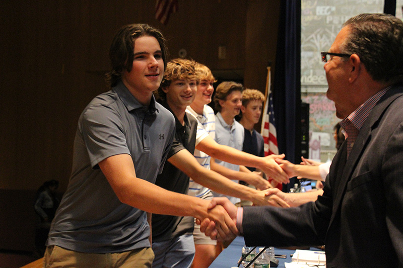 A line of high school boys shakes hands with adults on a stage.