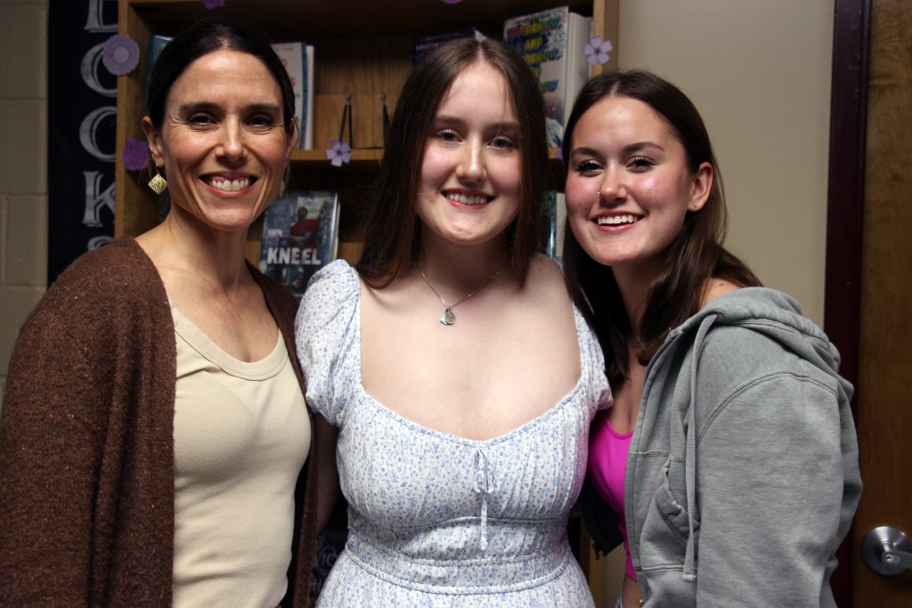 A young woman in a light colored dress, stands with two other women. All are smiling.