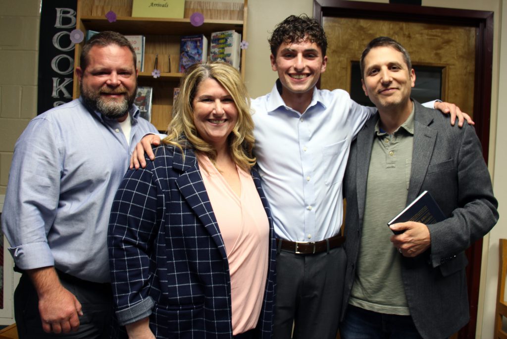 Four adults. A man on the left in a blue shirt, woman next to him with long blonde hair wearing a peach shirt and navy checked jacket, a high school senior with short curly dark hair, long sleeve button-down shirt, and a man on the right in a green jacket and shirt. He is holding a small book. All are smiling.