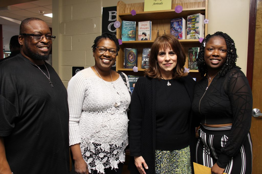 Four people stand togtether. A man on the right, with glasses wearing a black shirt, a woman next to him with short dark hair wearing a print skirt and white shirt, a woman wearing a black shirt and black print pants, and a high school senior wearing a black shirt and printed skirt. All are smiling.