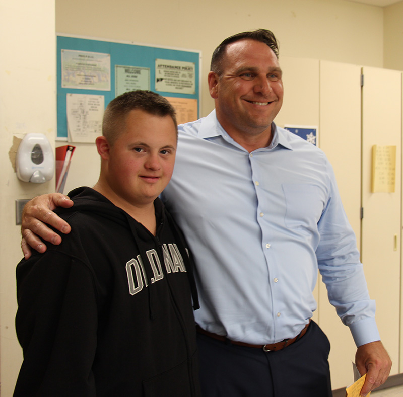 A young man wearing a blue Old Navy sweatshirt smiles as a man in a blue dress shirt puts his arm around him and smiles.