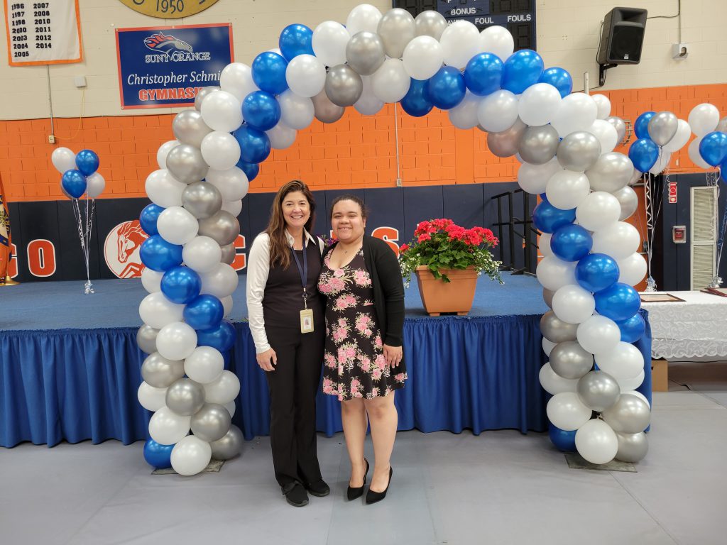 Under a blue, white and silver balloon arch stand two women. The woman on the left has shoulder length brown hair and is wearing black pants and shirt. The young woman on the right is a high school student wearing a flowered dress and dark sweater. Both are smiling.