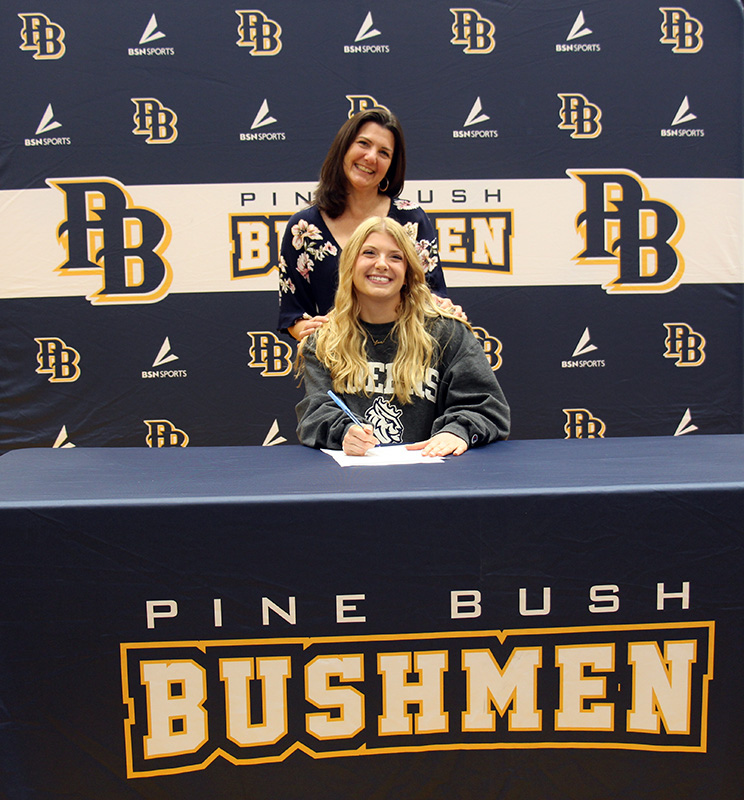 A high school senior girl sits at a table with a tablecloth that says Pine Bush Bushmen. She is smiling and a woman stands behind her holdin gher shoulders and smiling too. The backdrop says PB and Pine Bush Bushmen.