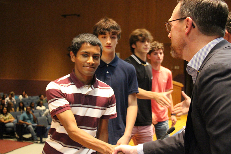 A high school student with short dark hair and a striped shirt shakes hands with a man on a stage.