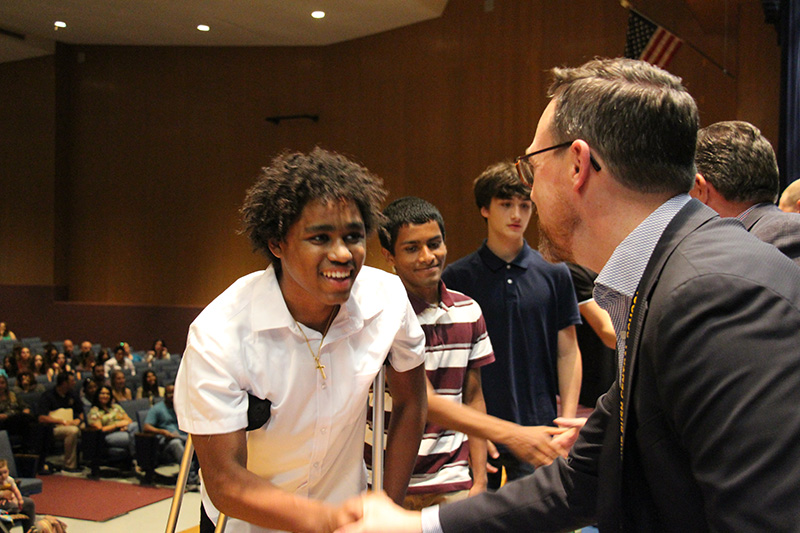 A high school young man in a white shirt smiles broadly and shakes hands with a man on a stage.