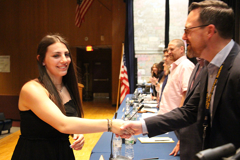 A young woman in a black dress and with long dark hair shakes hands with a man on a stage.