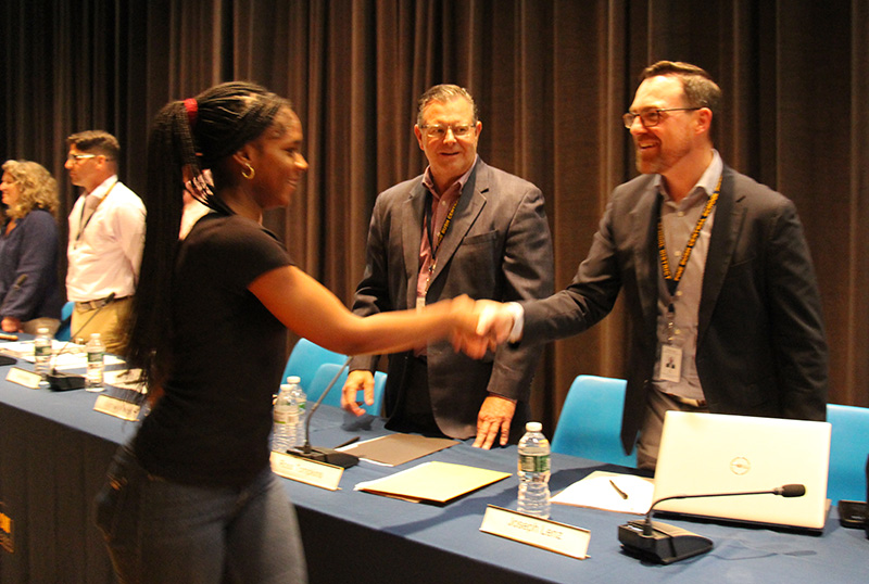 A young woman with her hair up in a ponytail shakes hands with a man at a table while another man looks on.