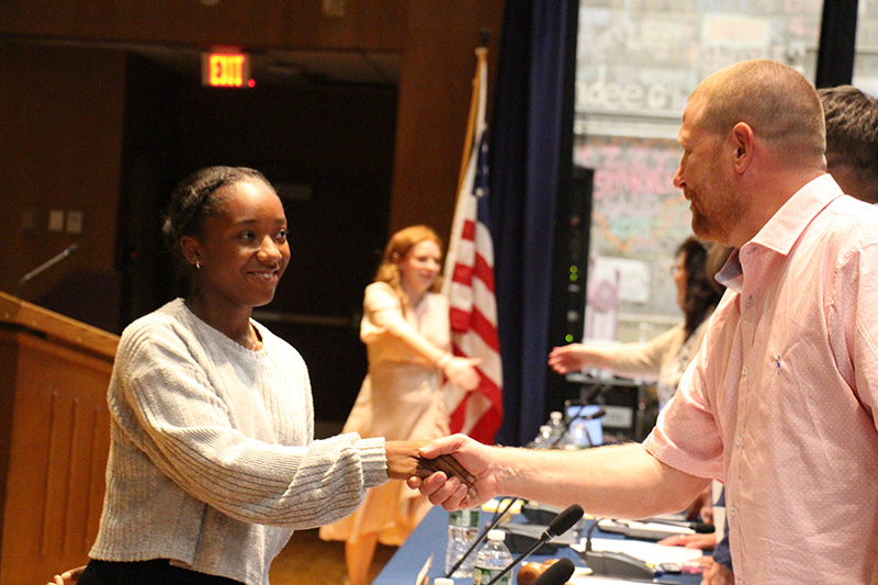 A high school girl wearing a white shirt smiles and shakes hands with a man on a stage.