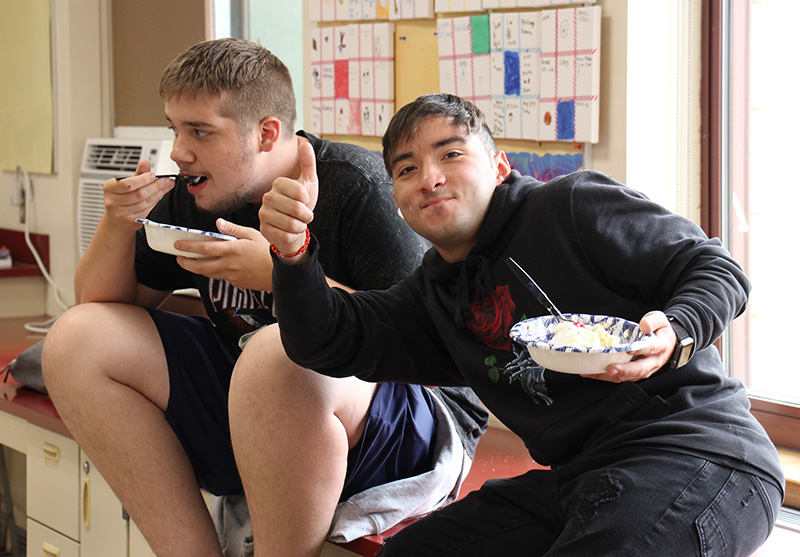 Two young men sit and eat bowls of ice cream. The young man on the right is giving a thumbs up.