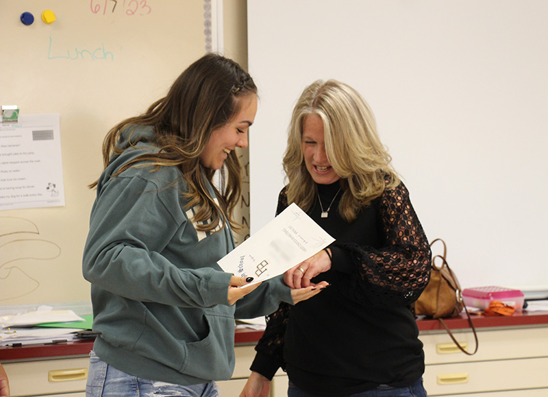 A young woman with long dark hair wearing a greensweatshirt accepts a certificate and pin from a woman with long blonde hair wearing a black shirt.