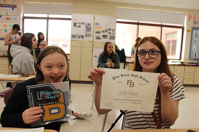 Two young women sit at desks and hold up items. One has a PB letter and the other a certificate. Both are smiling.