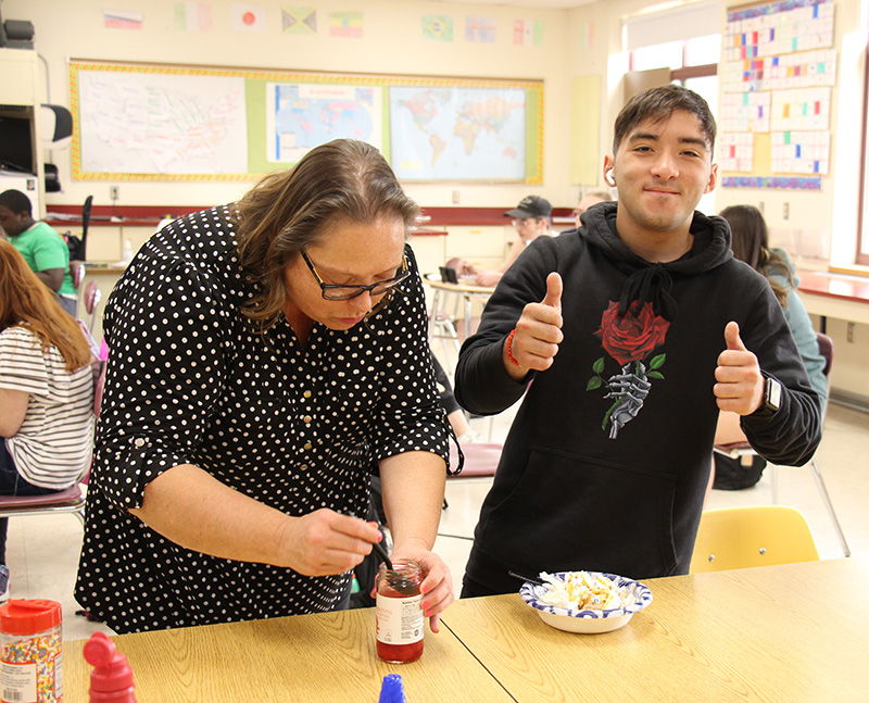 A woman gets a cherry  out of a jar to put on a dish of ice cream that is sitting next to it. A young man in a black sweatshirt gives two thumbs up.