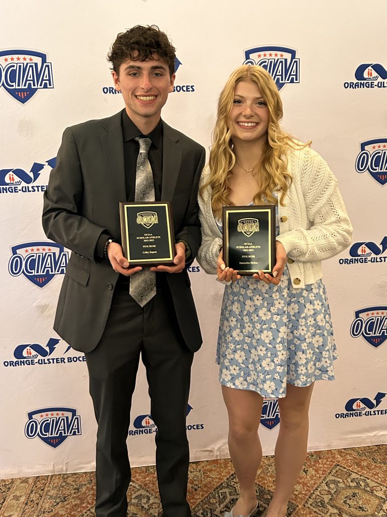 A young man on theleft is wearinga gray suit with a striped tie and holding a plaque. The  young woman on the right has long blonde hair and is wearing a blue and white dress and white sweater.The backdrop is white with blue writing and says OUBOCES. Both are smiling.