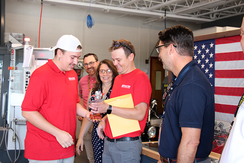 A high school senior, wearing a red shirt and white baseball cap is handed a small silver object he made, from a man also wearing a red shirt. There are three others looking on and smiling.