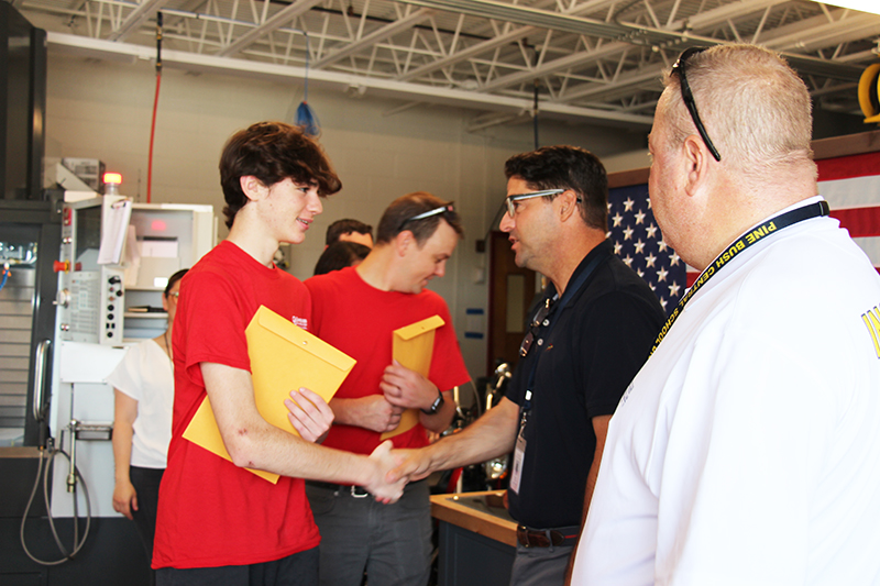 A high school student dressed in a red shirt holds a large manila envelop and shakes hands with a man with dark hair, glasses wearing a dark shirt.