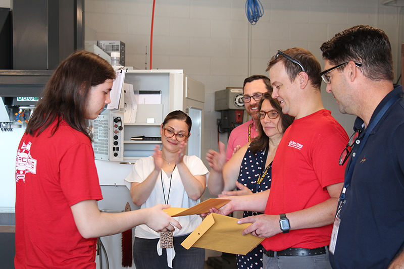 A high school young man shakes hands and accepts a manila envelope from a man also wearing a red shirt. Others are clapping as they look on.