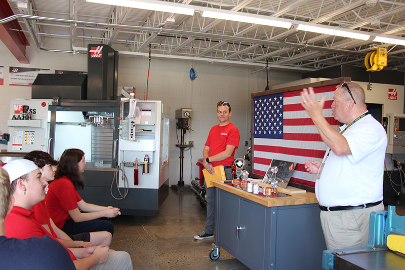 A man on the right wearing a white shirt speaks to a small group of high school students sitting in chairs. Another man, wearing a red shirt, stands to the left.