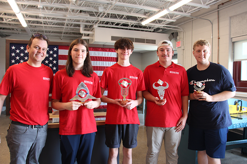 A man on the left and four high school students stand together. The young man on the right is wearing a dark blue shirt while the others are all wearing red shirts. The three in the middle hold projects they made from aluminum. All are smiling.