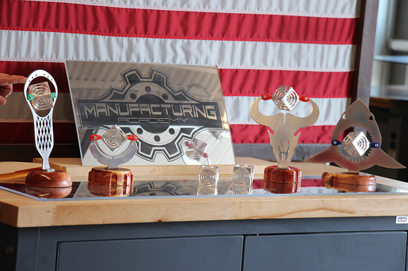 The background is an American flag. On a shelf in front are projects made by students out of aluminum.