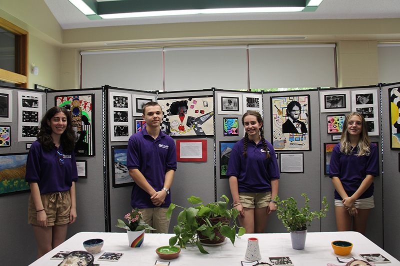 Four high school students - three girls and one boy - stand in front of a wall that contains artwork they produced.