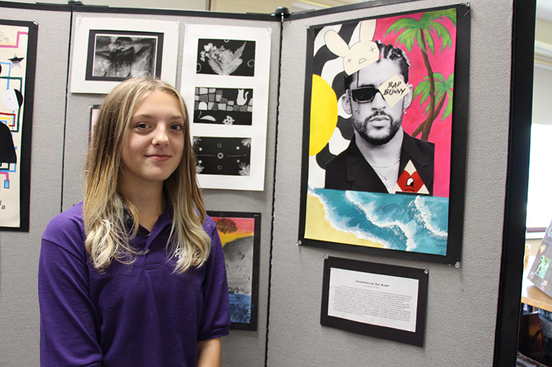 A young woman with long blonde hair stands in front of a picture she has worked on.