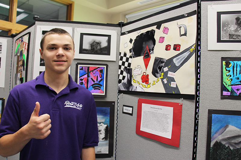 A young man with short dark hair gives thumbs up while standing in front of artwork he created.