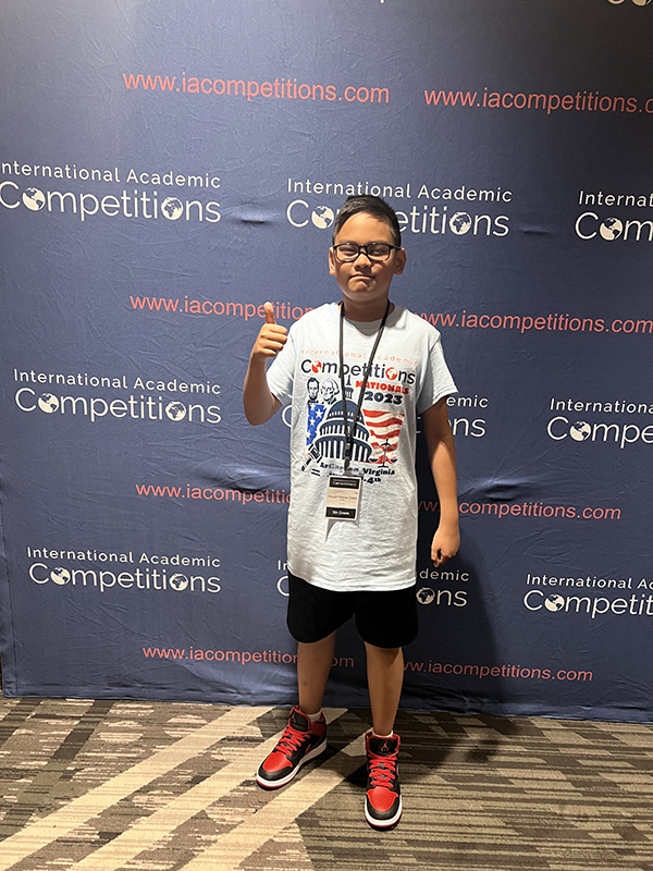 A fifth-grade boy with short dark hair and glasses wears a white tshirt with an American flag on it and gives a thumbs up. He is standing in front of a dark blue backgorund with International Competitions written on it.