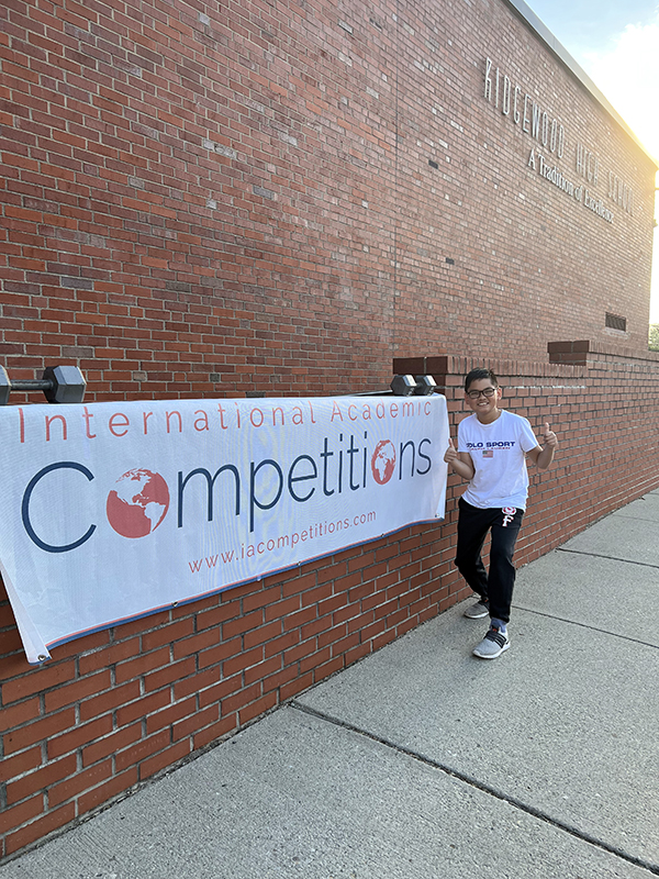 A long white banner hung on a brick wall which says International Competitions. A fifth-grade boy is at the far end of the banner giving a thumbs up.