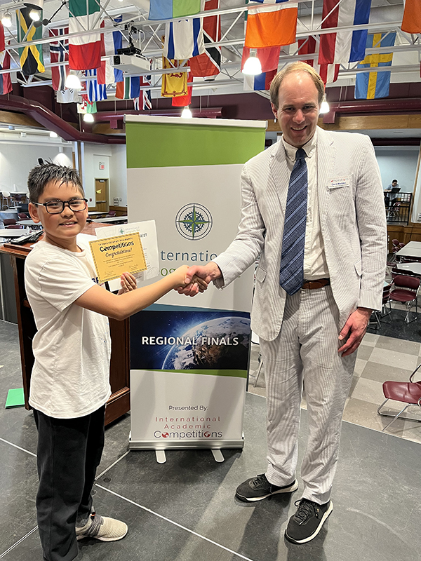 A fifth-grade boy with short dark hair and glasses shakes hands with a man wearing a light colored suit. Both are smiling.