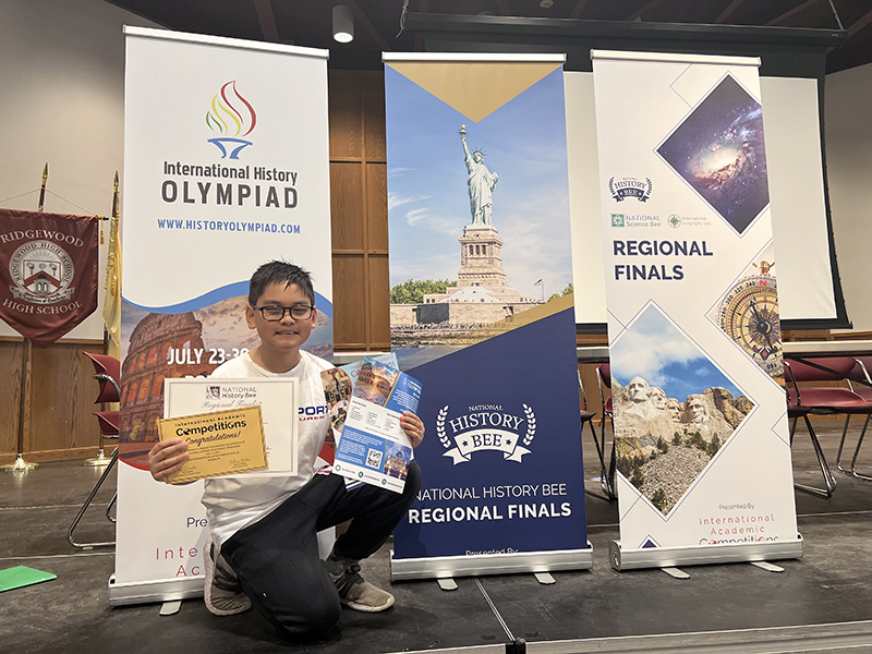 A fifth-grade boy stands in front of large vertical banners with information about academic competitions. He is crouching down holding certificates and smiling.