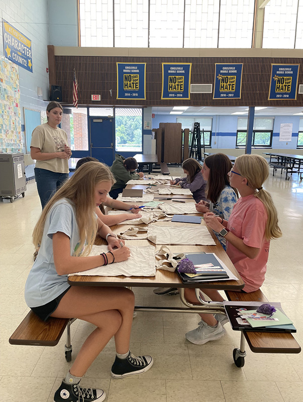 Five middle school girls sit at a table filling bags with items. A woman stands behind them .