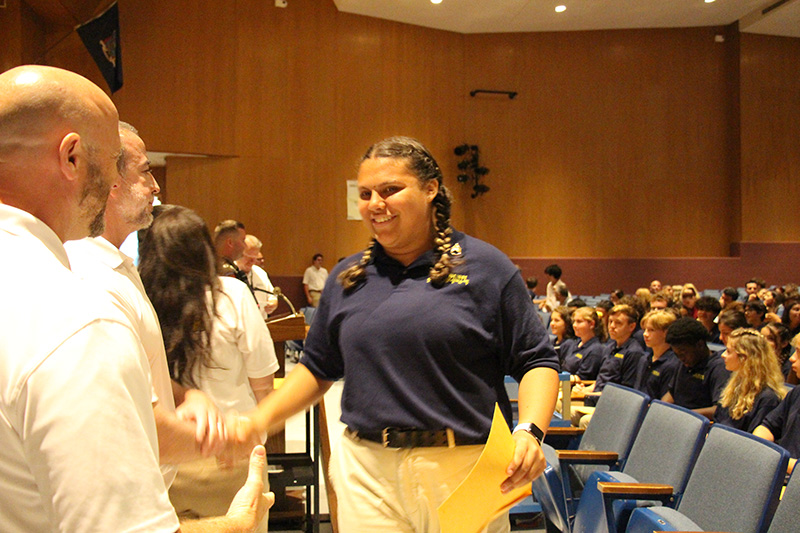 A high school aged girl wearing a dark blue polo shirt and tan pants, with two braids, smiles and shakes hands with a man as she accepts a certificate.