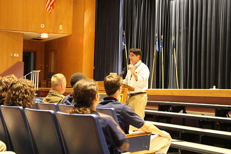 A man with dark hair, wearing a white shirt and tan pants, talks toa large group of people in an auditorium.