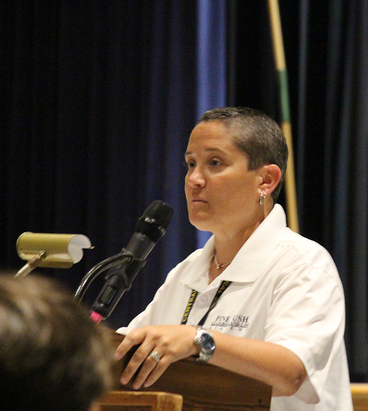 A close up photo of a woman wearing a white polo shirt speaking at a podium. She has short dark hair and is looking at the audience.