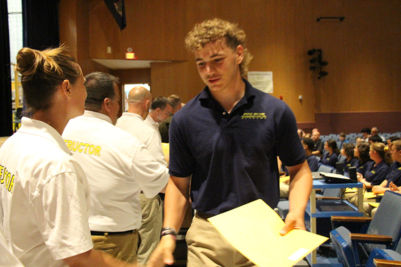 A high school aged boy wearing a dark blue polo shirt and tan pants, smiles and shakes hands with a woman as he accepts a certificate.