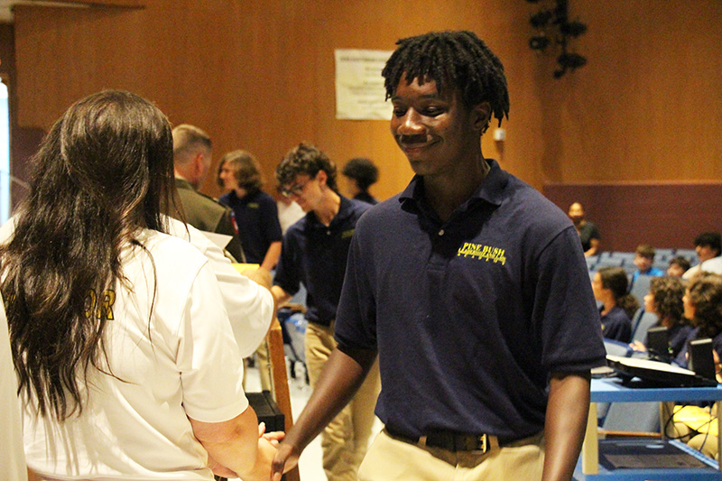 A high school aged boy wearing a dark blue polo shirt and tan pants, smiles and shakes hands with a woman as he accepts a certificate.