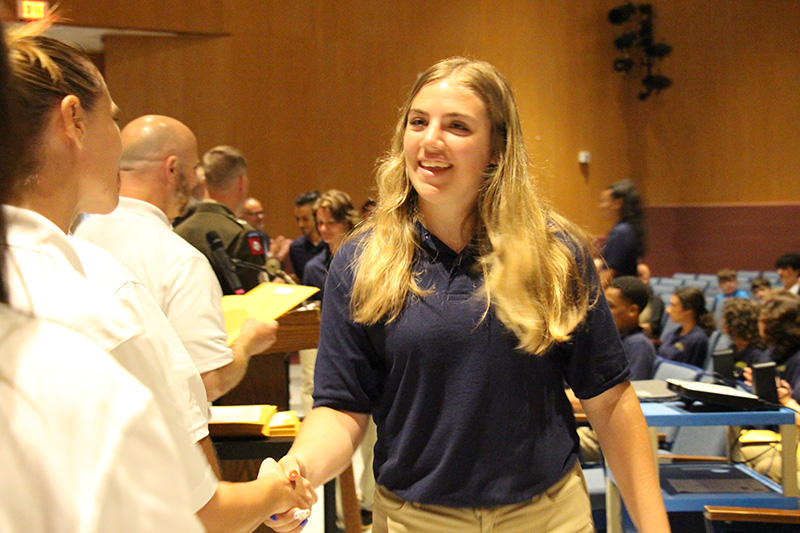 A high school age girl with long blonde hair, wearing a blue shirt and tan pants, shakes hands with an adult.