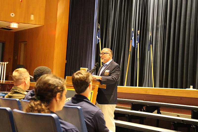 a man wearing a blue blazer and tan pants stands at a podium and talks to a large group of high school kids and parents.