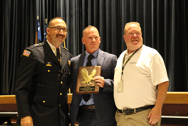 Three men stand together. The man in the center is wearing a suit and tie and holding a plaque. On the right is a man wearing a white polo shirt and tan pants. On the left is a man in a police uniform. They are all smiling.