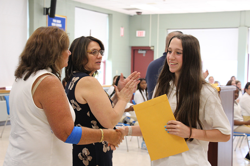 A young woman with long brown hair, wearing a white polo shirt, shakes hands with a woman after receiving a certificate.