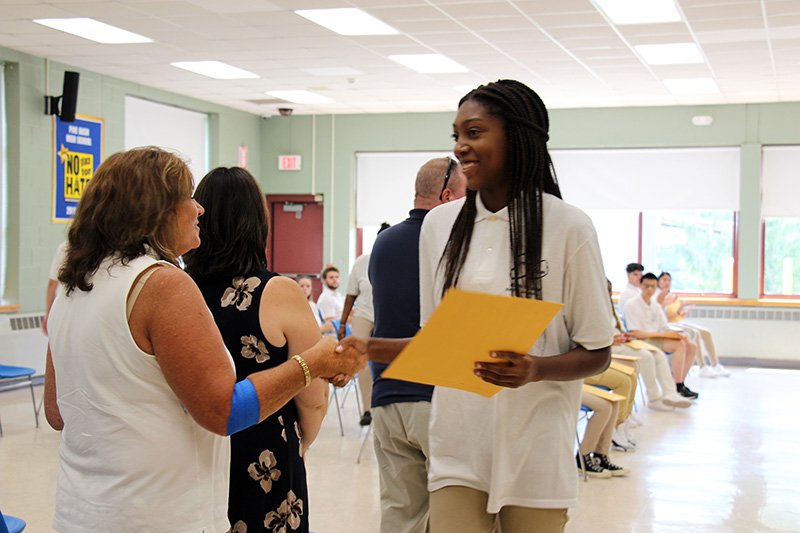 A young woman with long dark braids smiles as she shakes hands with a woman after receiving a certificate.